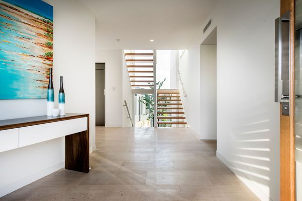 Bright contemporary residential entry hall with a floating white console and dark timber supports, colourful abstract artwork, decorative vases, and a sculptural open-riser timber staircase. Natural light enters through tall glazing and casts linear shadows across the pale walls and large-format stone or concrete-look floor.