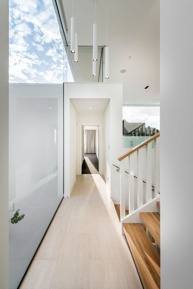 Bright contemporary residential hallway and stair landing with a double-height glazed wall, white walls and ceiling, pale large-format tiled flooring, and a timber stair with white balustrade and timber handrail. Slim white pendant lights hang above the circulation space, and a corridor leads toward a curtained room in the background.