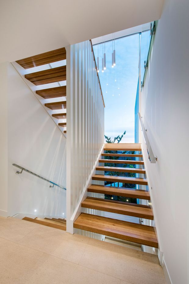 Bright contemporary stairway with open-riser timber treads, white walls, a vertical white slatted balustrade, and a glass balustrade along the adjacent stair flight. A large glazed opening brings in daylight and views of trees, while slim pendant lights hang in the stair void. The landing floor is finished in a light neutral stone or tile.