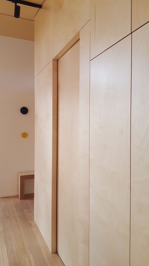 A narrow, minimalist interior passage dominated by pale timber or plywood wall panelling and flush concealed cabinetry or doors. The light timber floor continues through the space, with a simple white wall, small circular wall lights in black and mustard yellow, a timber-framed opening or bench at the far end, and a black ceiling track light. The image emphasises seamless joinery, clean lines, and warm natural materials.
