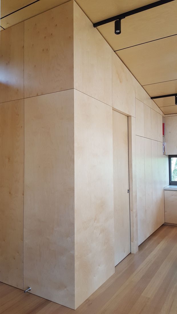 A narrow, minimalist interior corridor or passage lined with pale natural timber plywood wall panels and integrated cabinetry. A flush timber door is concealed within the wall, while the ceiling uses matching paneling with exposed black track lighting. Timber flooring continues through the space toward a small kitchen or built-in joinery area and window at the far end.