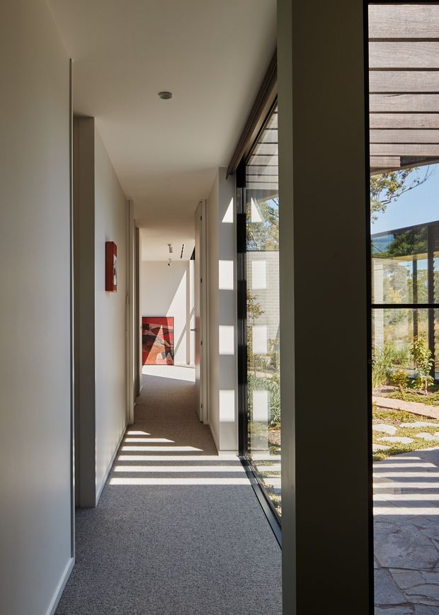 A long, narrow residential hallway with white walls, a smooth white ceiling, grey carpet, and simple flush doors. Full-height glazed panels with dark slender frames run along the right side, creating strong linear sunlight and shadow patterns across the floor and revealing a landscaped courtyard outside. A colourful abstract artwork anchors the far end of the corridor. The space has a restrained, contemporary architectural character with abundant natural light.
