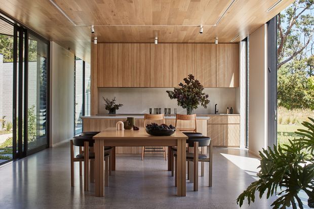 A bright, open-plan dining area with a timber-lined ceiling and full-height glazing overlooking landscaped grounds. A simple timber dining table with upholstered chairs sits in front of a wall of warm wood cabinetry and a streamlined kitchen or servery zone. The polished grey floor, neutral finishes, abundant daylight, and restrained furniture create a calm contemporary interior.