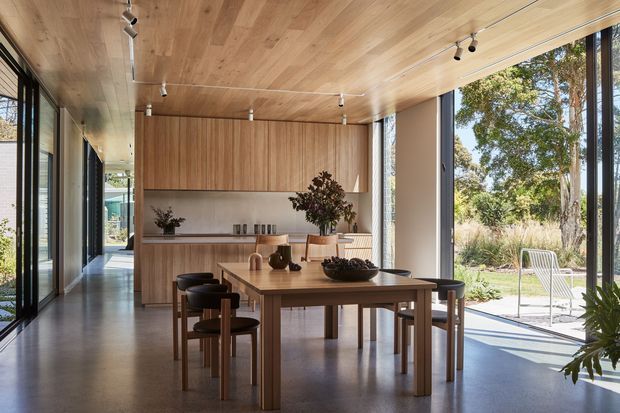 A bright, open-plan dining area with a long timber dining table and matching chairs and stools. Floor-to-ceiling glazing connects the room to a landscaped outdoor setting, while a full-height timber ceiling and built-in timber cabinetry create a cohesive natural-material interior. Minimal track lighting and restrained decor give the space a refined, contemporary character.