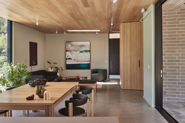 A refined open-plan interior viewed from a dining area, with a large solid timber dining table and black stools in the foreground, a lounge with dark sofas and armchairs beyond, and a timber-clad ceiling. Floor-to-ceiling glazing brings in greenery, while warm wood cabinetry, a polished concrete floor, neutral walls, track lighting, and a dark brick feature wall create a contemporary, natural-toned setting.