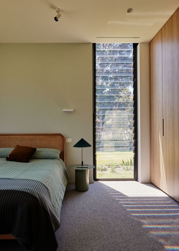 A calm, contemporary bedroom with a partially visible bed featuring a woven timber-toned headboard, soft green bedding and a dark accent cushion. The room has pale sage walls, a full-height black-framed louvred window or door bringing in dappled daylight, a full-height warm timber wardrobe wall, and a small round bedside table with a sculptural dark green lamp. The carpeted floor and restrained material palette create a warm, refined atmosphere.