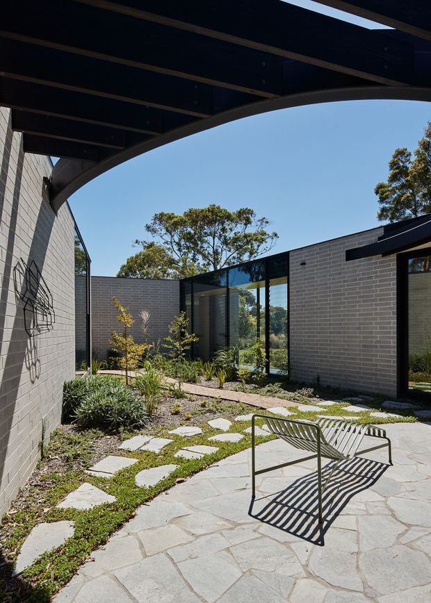 A sunlit modern courtyard enclosed by dark grey brick walls and extensive floor-to-ceiling glazing. Irregular pale stone paving forms a patio and stepping-stone path through a landscaped garden, with a minimalist metal bench in the foreground. A dramatic dark timber or metal overhead canopy with a curved edge frames the courtyard, while mature trees are visible beyond the walls.
