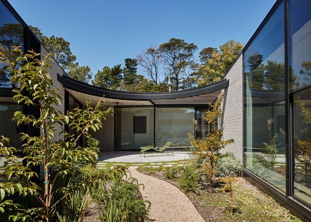 A contemporary single-storey courtyard house arranged around a landscaped central garden. Dark-framed floor-to-ceiling glazing and pale textured masonry walls sit beneath a distinctive curved roof edge, with a winding gravel path, native-style planting, and a small outdoor lounge area in the courtyard.