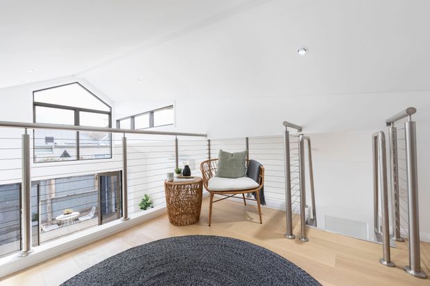 Bright upper-level landing or hallway overlooking a lower-level void through large windows and horizontal cable balustrades. The space has pale timber flooring, white walls and ceiling, a dark round rug, a woven side table, and a single upholstered timber-framed chair. A contemporary stainless-steel cable stair railing and open stairwell are visible on the right, with neighbouring building rooftops seen outside.
