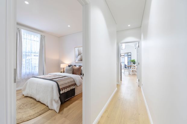 Bright contemporary hallway with pale timber flooring and white walls, looking toward a light-filled dining or living area. An open doorway on the left reveals a styled bedroom with a neatly made bed, upholstered headboard, bedside lamp, artwork, curtains and a round rug.