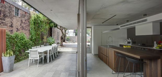 A contemporary open-plan kitchen opening directly onto a sheltered paved courtyard. The kitchen features dark wood-grain cabinetry, a long island with a dark benchtop, integrated appliances, and bar stools. Large glazed sliding doors create a strong connection between the interior and the outdoor dining area, which has a white table and chairs beside planted brick boundary walls.
