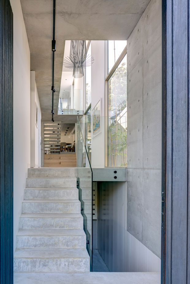 A tall, narrow interior stair hall with raw concrete stairs, walls and ceiling, a glass balustrade, and large vertical glazing. The stair connects multiple levels, with a dining area visible beyond and a sculptural linear pendant suspended in the double-height void. Dark exposed services and blue-grey door or wall framing add contrast to the pale concrete palette.