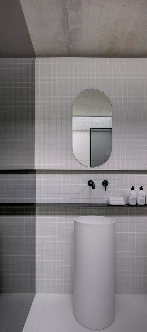 A minimalist contemporary bathroom featuring fine-grid light grey and dark grey wall tiles, a concrete-look upper wall and ceiling, a sculptural cylindrical white basin, matte black wall-mounted tapware, a slim dark benchtop, and an arched capsule-shaped mirror. Folded white towels and dispensers sit on the vanity.