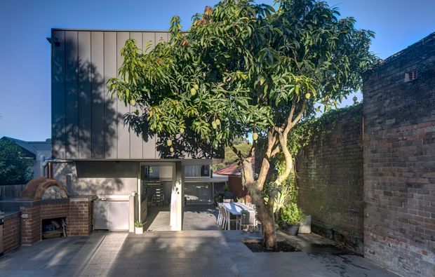 A contemporary rear courtyard or outdoor entertaining area viewed from outside, featuring a compact dark-clad two-storey extension with large glazing, a paved terrace, built-in brick barbecue and bench at left, outdoor dining beneath a mature tree, and an older exposed-brick boundary wall at right.