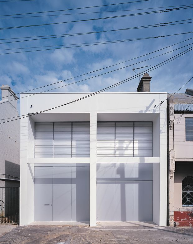 Front exterior view of a narrow contemporary urban building with a crisp white, rectilinear facade. The two-storey frontage has large upper-level windows fitted with horizontal metal louvres or blinds, recessed lower-level entry and garage-like doors, and a flat roofline. Adjacent older masonry buildings, overhead utility wires, a chimney, and strong sunlight with shadows are visible.