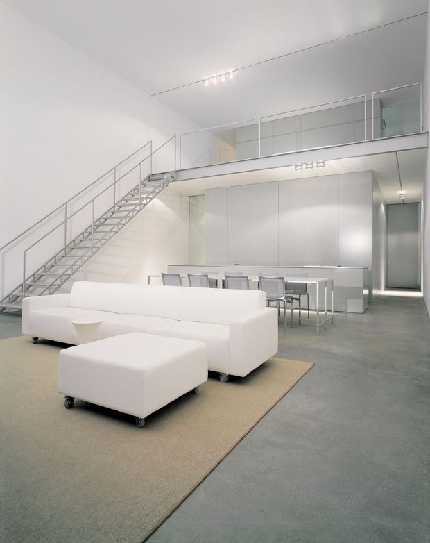 A bright, minimalist open-plan interior combining a living room and kitchen/dining area. White modular sofas and a large ottoman sit on a neutral rug in the foreground, while a compact white kitchen with a long island and stools occupies the rear. The space has a polished grey concrete floor, white walls and ceiling, extensive built-in cabinetry, a glass-balustraded mezzanine, and an exposed metal stair with slender railings.