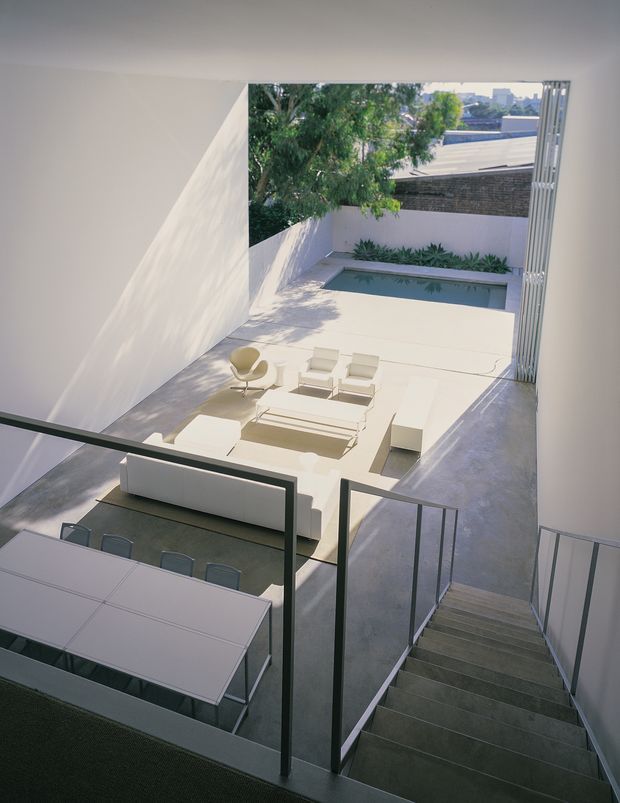 View from an upper-level stair landing down into a minimalist open-plan living and dining area, with a large white dining table in the foreground, white sectional seating, and a sculptural lounge chair. The room opens directly onto a bright paved courtyard terrace with a narrow rectangular pool, linear garden planting, high white walls, and an adjacent dark brick boundary wall. A glazed or open vertical-slatted facade frames the right side, while dark metal stair and balustrade elements contrast with the pale finishes.