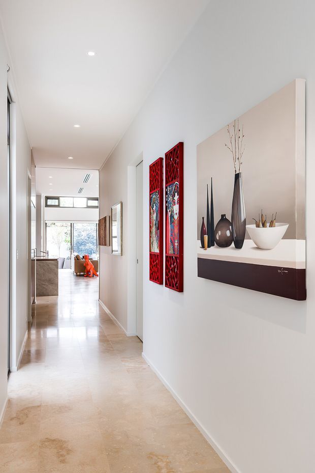 A bright, narrow residential hallway with white walls and ceiling, recessed downlights, and a pale polished stone or concrete-look floor. A floating dark wood display shelf with a white top holds sculptural vases and branches on the right wall, accompanied by two vivid red decorative wall panels. The corridor leads toward a glazed living area with a glimpse of seating and a kitchen or joinery beyond.