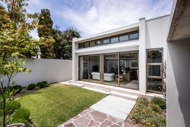 A contemporary single-storey house viewed from a landscaped courtyard garden. The white rendered facade features extensive timber-framed glazing, a high clerestory window band, and a sheltered glass entry door. A paved path combines large light concrete slabs with irregular natural stone, leading through a neat lawn and low planting to the living area visible inside.