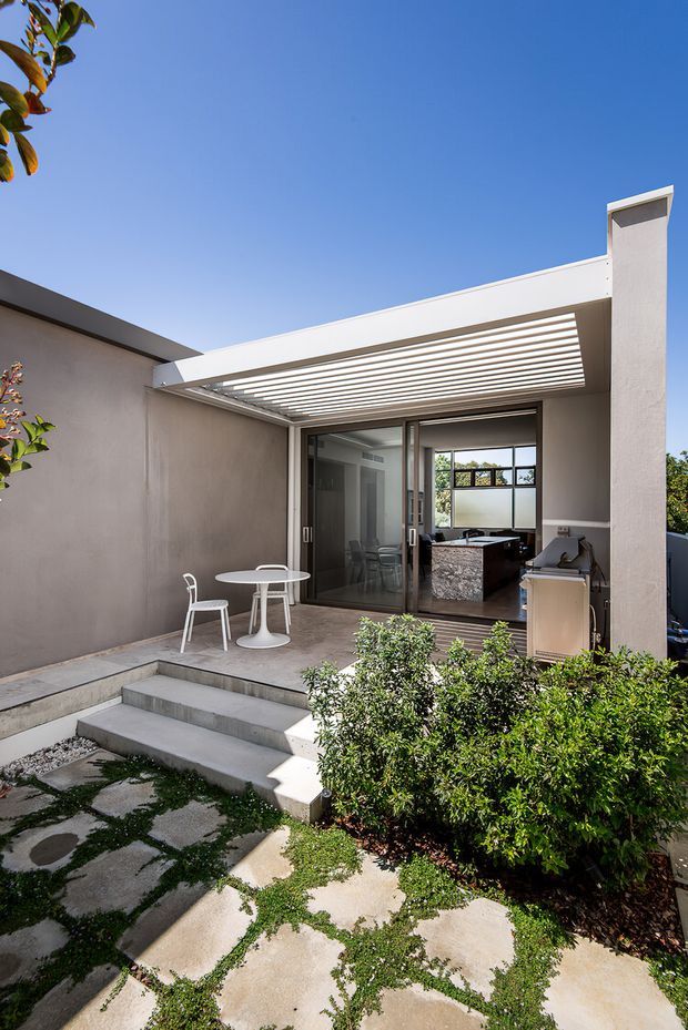 A contemporary courtyard patio viewed from the garden, with pale rendered walls, broad concrete steps, irregular concrete paving, dense shrubs, and a white pergola with horizontal slatted shading over a small outdoor dining setting. Large sliding glass doors open into a bright interior that appears to contain a kitchen or living area with a dark stone island or counter. The scene is sunlit with a clean, minimalist architectural character.