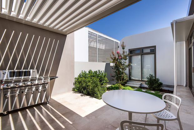 A bright, contemporary courtyard entertaining area with a stainless-steel outdoor barbecue beneath a louvred overhead canopy. The paved space includes a round outdoor dining table and chairs, landscaped garden beds, a feature tree, and a modern white rendered wall with glazed doors and screening.