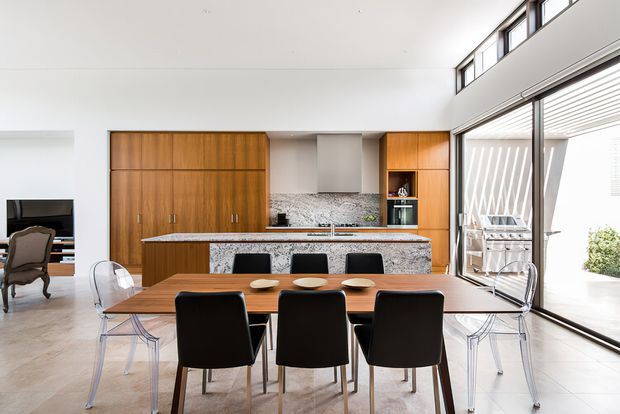 Bright open-plan dining and kitchen interior with a long timber dining table surrounded by black and transparent chairs. The kitchen features extensive warm timber cabinetry, a pale stone island and benchtop, patterned stone backsplash, integrated appliances, and a compact wine or display niche. Full-height glazed sliding doors open to a covered outdoor area, while high-level clerestory windows bring in additional daylight. A small living area is partially visible to the left.