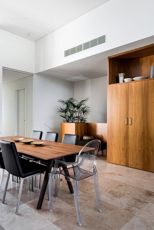 Bright contemporary dining area with a large rectangular timber dining table, black and transparent chairs, pale stone-look tiled flooring, white walls and a tall built-in timber storage unit with open display shelving. A potted palm adds greenery, while a high linear air-conditioning grille emphasizes the clean modern interior.