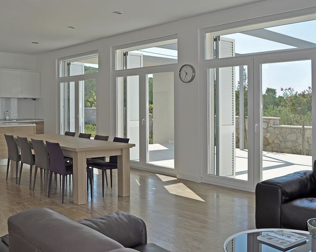 Bright open-plan interior combining a dining area and living room, with a large timber dining table and upholstered chairs in the foreground and a dark leather sofa with round coffee table at right. Full-height glazed doors and windows open toward a sunlit terrace, with white walls, recessed ceiling lights, pale timber flooring, and minimal white kitchen cabinetry. The space has a clean, contemporary design with abundant natural light.