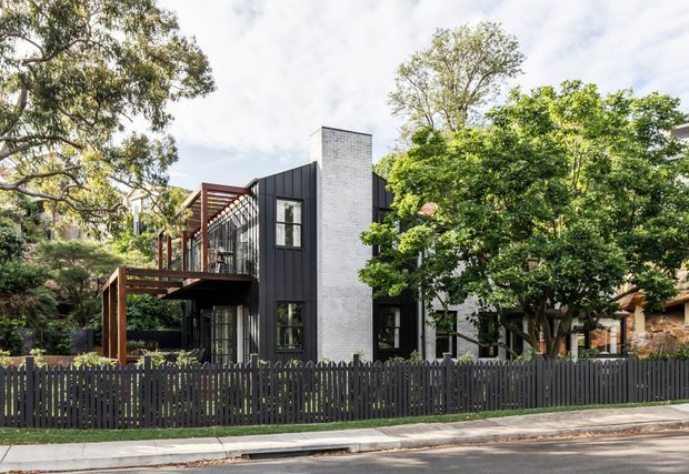 Street-facing view of a contemporary multi-level residential house set among mature trees. The facade combines dark vertical cladding, a prominent light brick chimney or feature wall, extensive glazing, and a timber-framed upper deck or pergola. A dark vertical-slat fence defines the front boundary, with neighbouring homes partially visible through the landscaping.