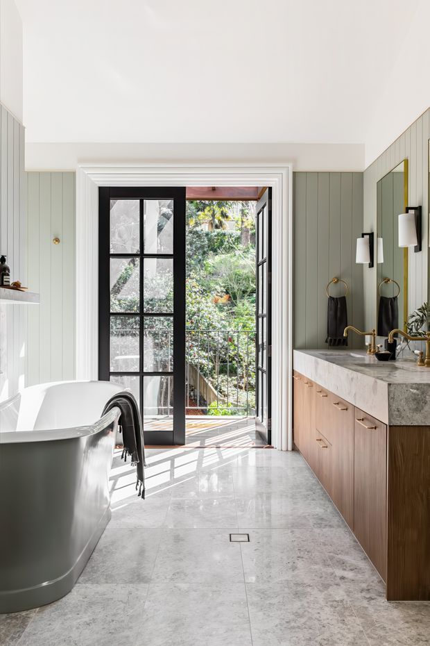 Bright, refined bathroom with a freestanding white bathtub, large-format pale stone-look floor tiles, and a timber vanity with a thick marble or stone benchtop. Vertical sage-grey wall panelling surrounds the room, while black steel-framed glazed doors open to a leafy outdoor balcony or garden. Brass tapware, wall sconces, and dark towels provide restrained accents.