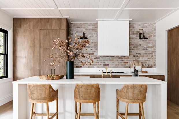 Bright kitchen featuring a large white island with three woven rattan bar stools, warm timber cabinetry, a brick feature backsplash, white panelled cabinetry, brass tapware, and a white-painted timber-lined ceiling. Black-framed glazing and a timber door add contrast, while natural decorative branches bring an organic accent.