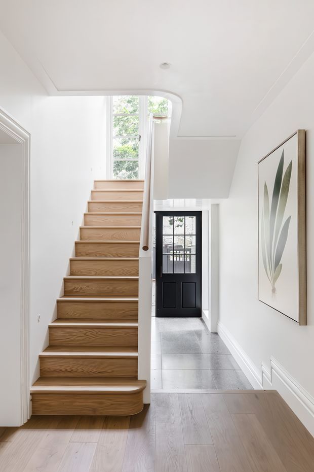 Bright residential hallway and stairway with a light timber staircase, white walls and ceiling, pale timber flooring, grey tiled entry flooring, and a black glazed front door. A large botanical artwork hangs on the wall, while daylight enters through tall windows at the stair landing and front entry.