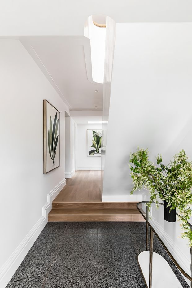 Bright, refined residential stairway and landing with white walls, decorative wall mouldings, framed botanical artwork, warm timber steps and hallway flooring, and a dark terrazzo-like lower floor. A curved glazed balustrade and indoor greenery frame the foreground. A tall, sculptural skylight or clerestory opening brings daylight into the stairwell.
