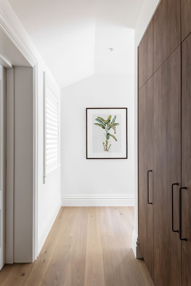 Bright, narrow residential hallway with pale timber flooring, white walls and decorative trim, plantation shutters on the left, and a full-height bank of dark timber built-in cabinetry with black handles on the right. A botanical print in a dark frame provides a subtle feature on the end wall. The space is clean, refined and naturally warm with contemporary detailing.