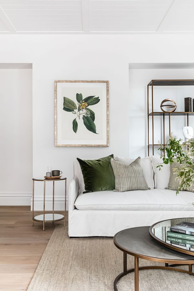 Bright, refined living room with a white slipcovered sofa, layered neutral and deep green cushions, a round dark coffee table, and a slim open metal shelving unit. The room features white walls and a white panelled ceiling, warm timber flooring, a large neutral rug, framed botanical artwork, and indoor greenery. The styling is calm and contemporary with subtle classic and natural accents.