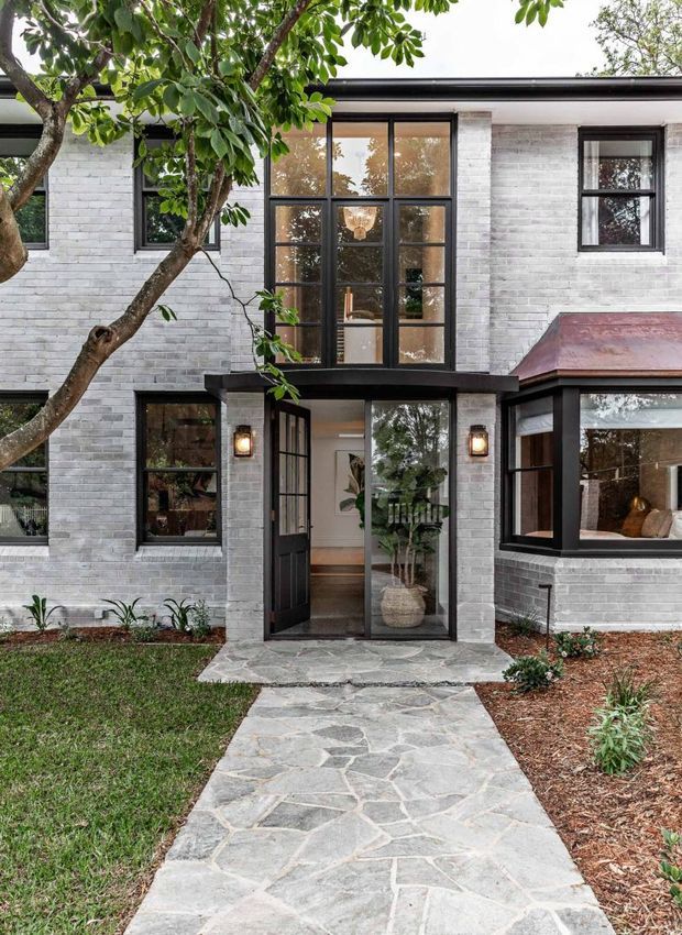 Front exterior view of a two-storey residence with white-painted brick walls, black-framed windows and glazed entry doors, a projecting bay window, and a dark low-profile roof edge. A mature tree and planted garden beds frame the irregular natural-stone front path leading to the entrance. Warm entry sconces and visible indoor furnishings add a welcoming character.