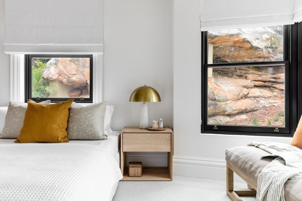 Bright, refined bedroom interior with a neatly made bed, white bedding, a mustard accent cushion, pale upholstered cushions, a timber bedside table, and a brass dome table lamp. White Roman blinds and black-framed windows overlook rocky terrain, while a cushioned timber bench sits beside the window. The room has a clean contemporary feel with neutral finishes and warm wood and brass accents.