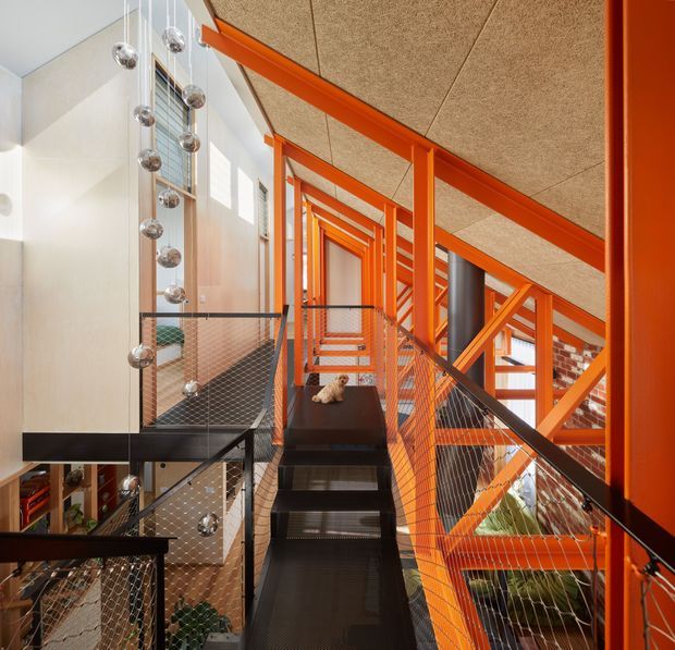 Interior view of a multi-level stairway and landing with a strong industrial character. Bright orange-painted steel beams and stair framing create a repeating structural rhythm, while black metal stair treads and wire-mesh balustrades overlook a lower level. The space has a textured acoustic-panel ceiling, exposed brickwork, extensive glazing, and a suspended installation of reflective spherical lights.