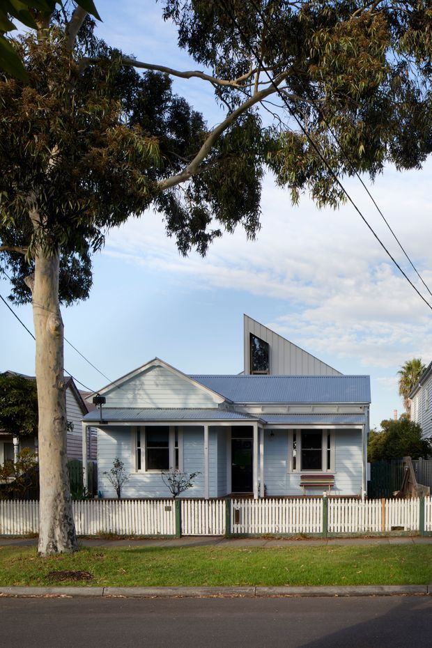 Street-facing exterior view of a compact single-storey weatherboard house with a gabled front roof, corrugated metal roofing, white picket fence, and a distinctive contemporary angled roof-volume extension rising behind the original cottage. Mature eucalyptus trees frame the property.