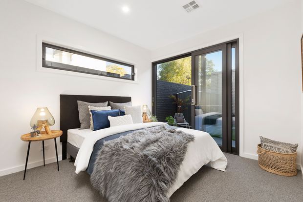 A bright, contemporary bedroom with a low upholstered dark headboard, layered white and grey bedding, blue and patterned accent cushions, and a large grey faux-fur throw. A small round timber side table with a sculptural lamp sits beside the bed. The room has pale walls, soft grey carpet, a narrow horizontal window, and full-height dark-framed glazed doors opening to a small outdoor area. A woven basket adds texture in the corner.