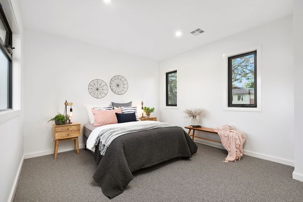 Bright, minimalist bedroom with white walls and ceiling, grey carpet, a centred bed dressed in dark grey and soft pink textiles, a timber bedside table, simple wall decor, and black-framed windows. The room has recessed ceiling lighting and a clean, contemporary fit-out.