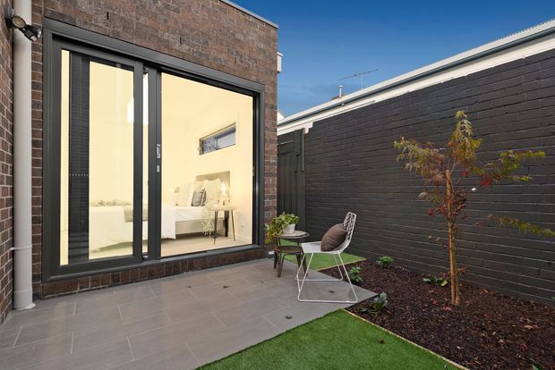A compact, enclosed courtyard beside a contemporary brick extension. Dark brick walls, charcoal-framed floor-to-ceiling glazing, grey tiled paving, a small outdoor seating arrangement, and a narrow landscaped garden create a private urban outdoor space. A bright bedroom is visible through the glass doors.
