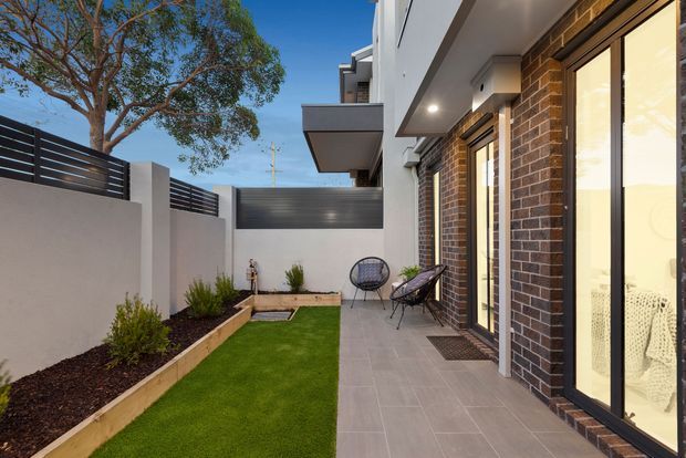 A modern residential courtyard or side garden beside a brick-clad home, featuring a narrow artificial-grass lawn, raised garden beds, rendered boundary walls with horizontal dark screening, and a tiled outdoor passage. Large glazed doors open onto the paved area, with a small outdoor seating setting near the house. The scene has clean contemporary detailing and a warm neutral palette.