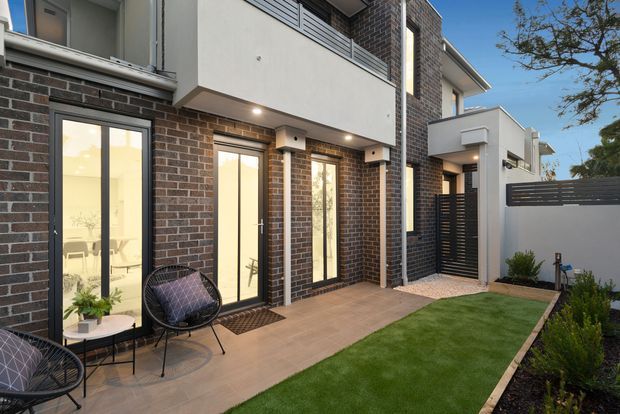 Contemporary multi-storey townhouse exterior with dark brick and white rendered walls, tall glazed doors, a tiled patio, landscaped lawn and a private side courtyard. Black metal screening and balcony balustrades add contrast, while warm interior lighting creates an inviting evening atmosphere.