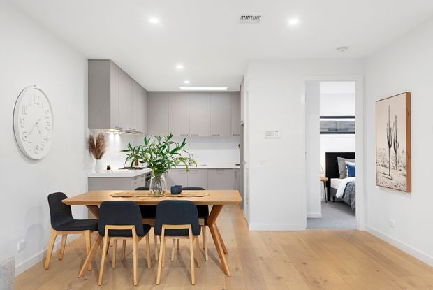 Bright open-plan dining and kitchen area with a light timber dining table and chairs in the foreground, a white and pale grey minimalist kitchen with integrated cabinetry behind, and a doorway leading to a bedroom. The room features pale timber flooring, white walls, recessed lighting, indoor plants, and understated wall art.