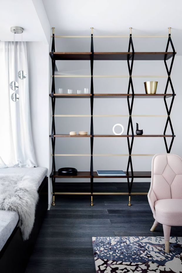 A refined bedroom corner featuring a tall open shelving unit with dark timber shelves, slim black geometric supports and brass-toned hardware. A pale pink upholstered accent chair sits on a patterned rug beside a dark bed with a soft grey throw. White curtains, a sculptural pendant light, dark timber flooring and carefully styled decor create a contemporary, polished interior.