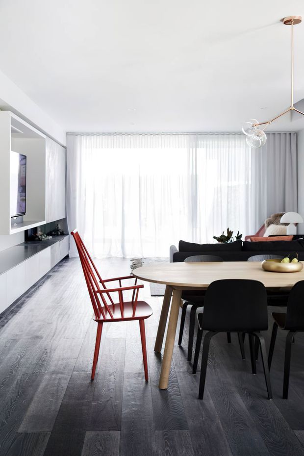 Bright open-plan dining and living room with floor-to-ceiling sheer curtains, dark timber flooring, white built-in media joinery, a light timber dining table, black dining chairs, and a distinctive red timber chair. A black sofa, floor lamp, pendant light, and indoor plant are visible in the living area.