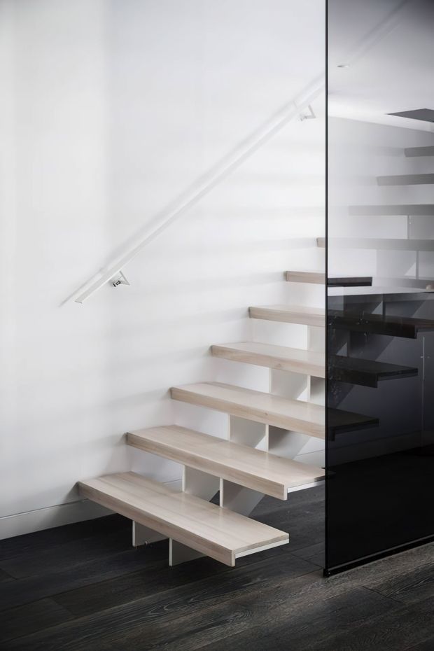 A minimalist floating staircase with pale timber treads and white supporting elements rises beside a glossy dark glass balustrade or partition. The stairwell has white walls, a slim white handrail, recessed lighting, and dark timber flooring, creating a crisp contemporary interior.
