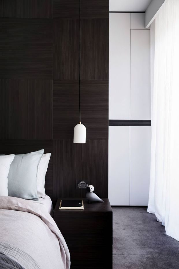 A refined contemporary bedroom corner featuring a bed with pale layered bedding, a dark vertically textured timber feature wall, matching dark bedside table, sculptural pendant light, and a sleek white built-in wardrobe. Floor-to-ceiling sheer curtains provide soft natural light, while grey carpet adds a muted neutral base.