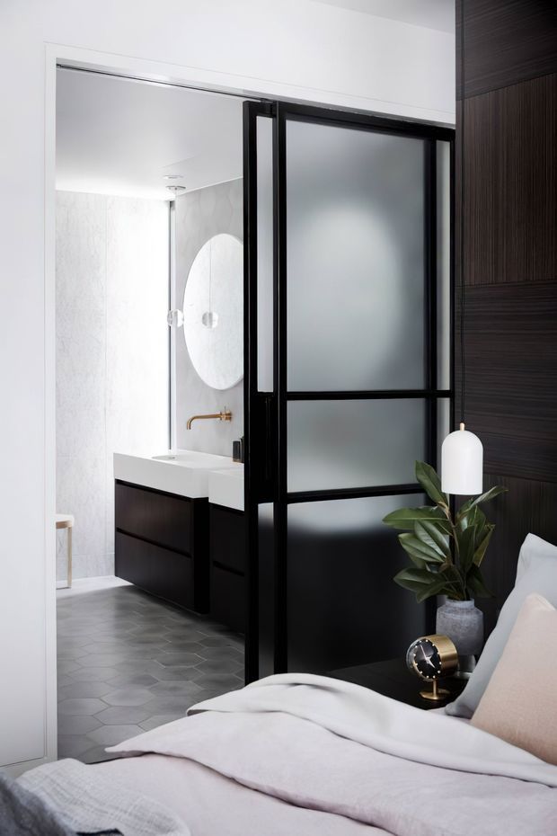 A refined bedroom looking through a black-framed frosted-glass partition into an adjoining bathroom. The bedroom features pale bedding, a dark timber wall, a bedside table and plant, while the bathroom has a dark floating vanity, white benchtop, pale wall tiles, a round mirror and brass tapware. The design uses strong black-and-white contrast with warm metallic accents.